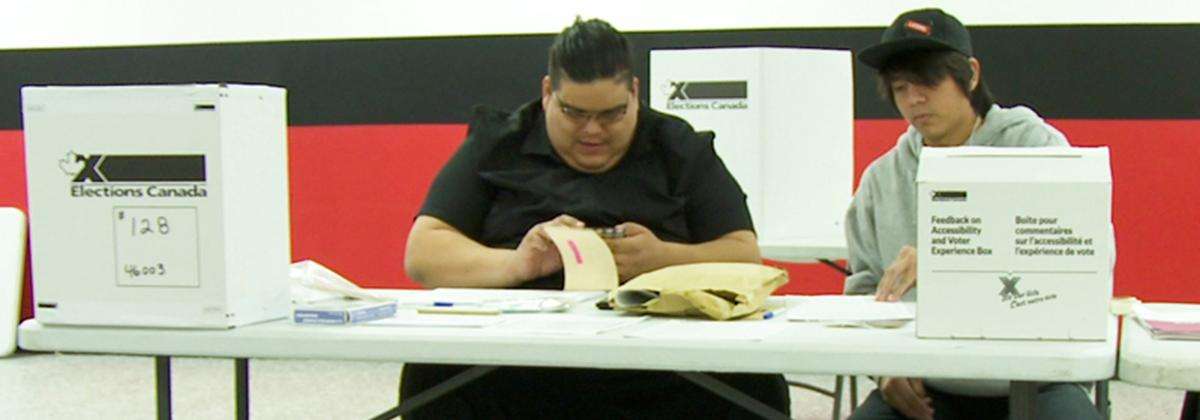 GE44 - Working at an election blog In a polling station, two poll workers are seated at a table with election materials.