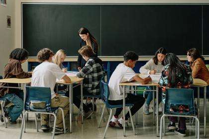 A classroom. There are two groups of students. Their desks are placed together to work as a team. A teacher is walking between the two groups of students.