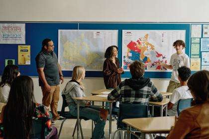 In a classroom, maps are displayed on the board. A teacher and two students are at the front of the classroom, next to the maps. They are speaking to the group of students.