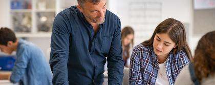 In a classroom, students write at their desks. A teacher and a student look at an assignment together.