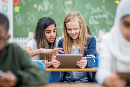 In a classroom, students are seated at their desks. Two students are doing a learning activity using a tablet.