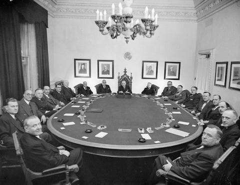 Black and white photo of a cabinet meeting. A group of parliamentarians are seated at a large oval table. 