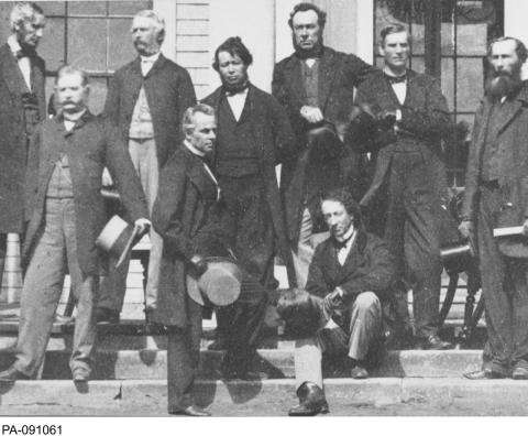 Black and white photograph showing a group of men, including Sir John A. MacDonald, gathered on steps holding their top hats.  