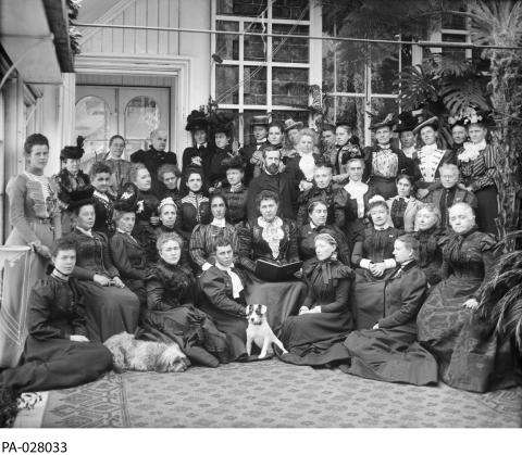 Black and white photograph showing a large group of women, positioned in rows. Two dogs sit in front.