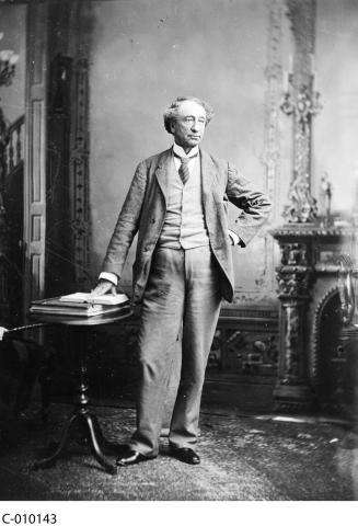 Black and white photograph of Sir John A. MacDonald standing in front of a fireplace, one hand on his hip, the other touching an open book on a small table beside him.  