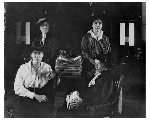 Black and white photograph showing four women seated around a table. On the table is a large stack of paper.