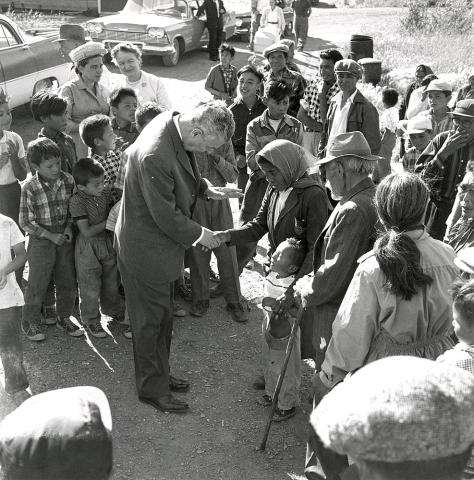 CE19-Women Black and white photograph showing Prime Minister John Diefenbaker shaking hands with a First Nations woman, amid a small crowd of people of all ages. The woman has her arm around a young child.