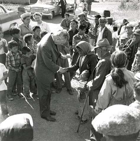 Black and white photograph showing Prime Minister John Diefenbaker shaking hands with a First Nations woman, amid a small crowd of people of all ages. The woman has her arm around a young child.