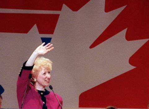 Photograph of Kim Campbell standing behind a microphone, smiling and waving.