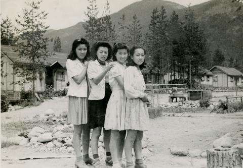 Black and white photograph of four young Japanese women standing in front of an internment camp, with their hands on one another's shoulders. Mountains are in the background.