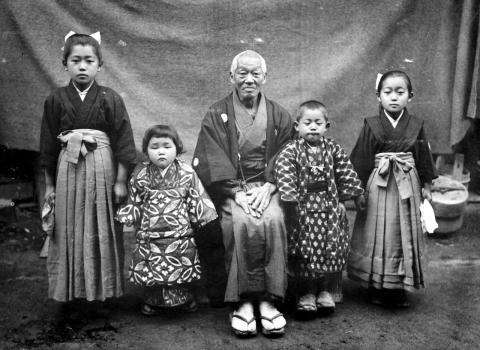 A black and white photograph of a Japanese family of five posing in traditional attire. An older gentleman sits in the middle, with two children on either side.