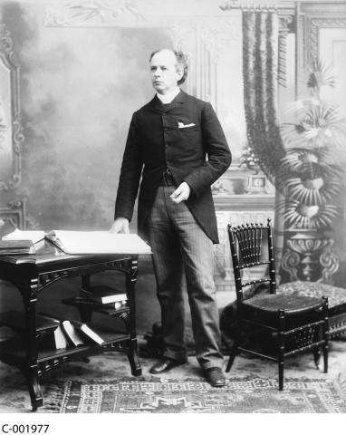 Black and white photograph of Wilfred Laurier, standing beside a table with his hand resting on a large open book.