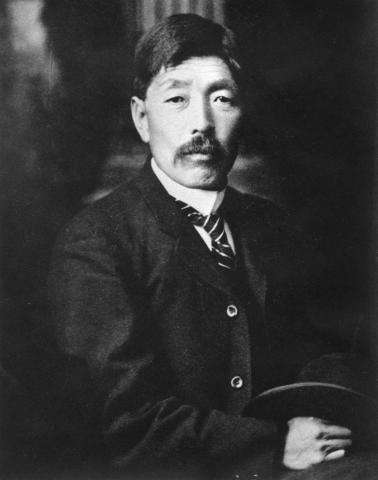 Black and white photograph of a mustachioed Japanese man, taken from the waist-up, holding his hat in his hands.