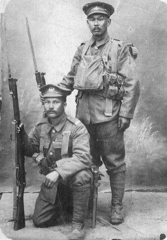 Black and white photograph of two Japanese men in military uniform, holding rifles at their sides. One man stands, and the other kneels on one knee in front of him.