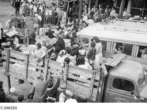 A black and white photograph showing many people of all ages being loaded into the back of a truck, with their luggage. Many others wait in line behind a rope barrier.