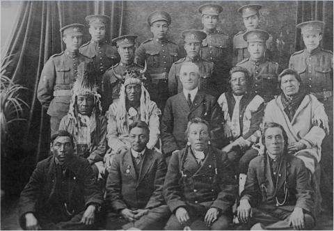 Black and white photo of the first time Indigenous men joined the Canadian Forces as soldiers.  First two rows: Indigenous men, some in traditional regalia. Last two rows: men in their soldier uniform.