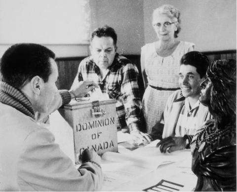Black and white photo in Hiawatha Council Hall on the occasion of a federal by-election. The first votes cast since the right to vote was extended to all status Indians were those of the Rice Lake Band near Peterborough, Ontario. From left to right: Lawrence Salleby; Chief Ralph Loucks, Deputy Returning Officer; Lucy Muskrat, Poll Clerk; Eldon Muskrat, Poll Constable.