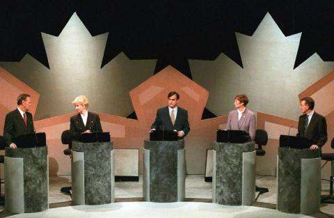 Picture of a televised leaders’ debate during the 1993 federal election. Five politicians are standing at podiums, in front a background of two large maple leaf cut-outs.