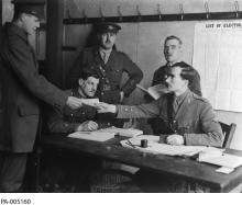 CE04-Youth Black and white photograph of a group of men wearing military uniforms, gathered around a table. One man gives a ballot to another man. On the wall is posted a paper that reads: “List of Electors.”
