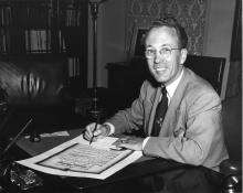 CE05-Youth Black and white photograph of Tommy Douglas sitting in his office, signing and official document and smiling at the camera.