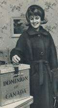 CE11-Women Black and white photograph of a young women placing her ballot in a metal ballot box, while smiling.
