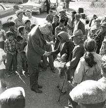 CE19-Women Black and white photograph showing Prime Minister John Diefenbaker shaking hands with a First Nations woman, amid a small crowd of people of all ages. The woman has her arm around a young child.