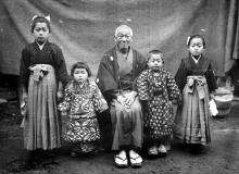 CE33-Japanese-alt 1 A black and white photograph of a Japanese family of five posing in traditional attire. An older gentleman sits in the middle, with two children on either side.